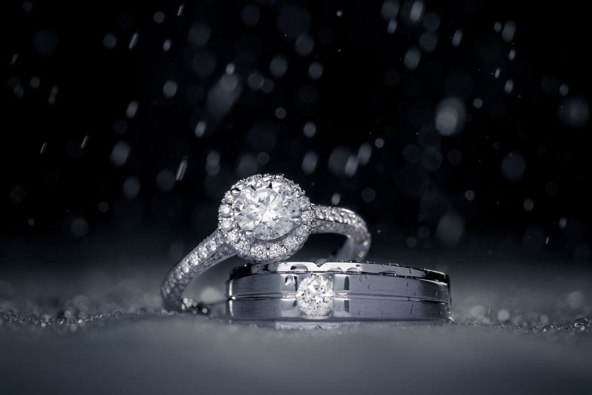 Diamond halo engagement ring and matching diamond wedding band in white metal with water droplets against a dark background